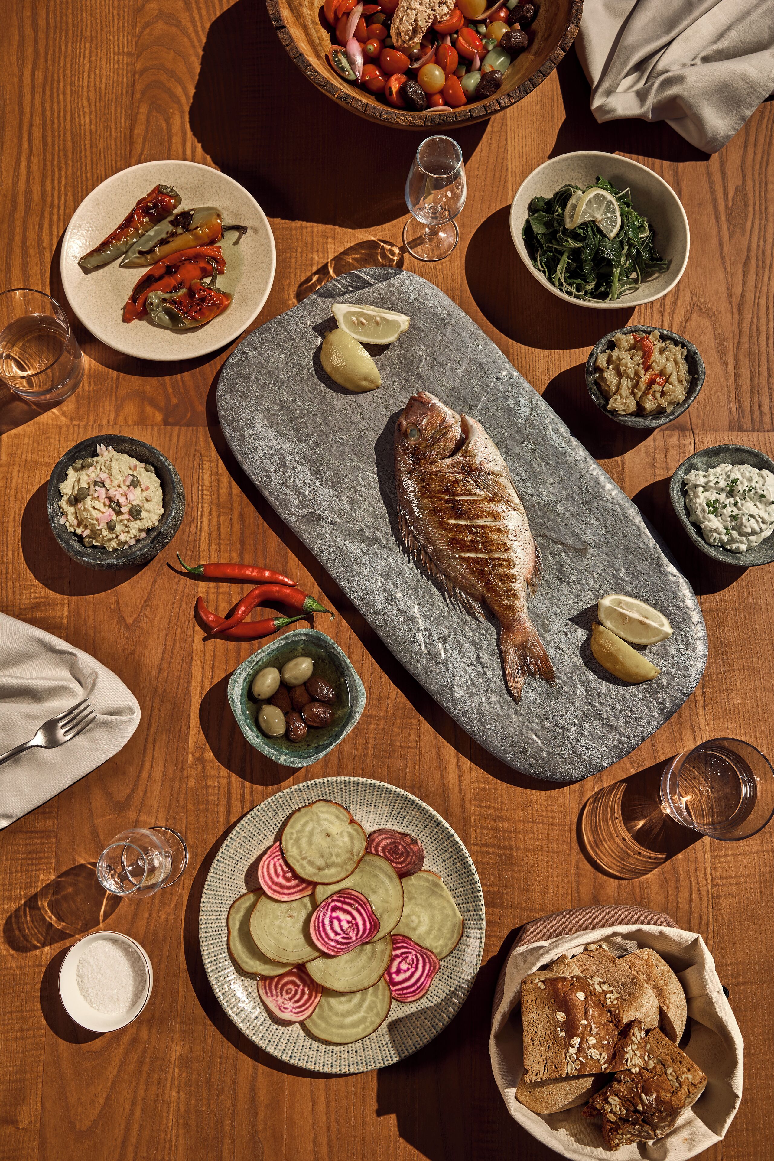 A wooden table with various dishes including a whole fish, olives, pickled vegetables, and bread.