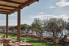 Un área de comedor al aire libre con mesas preparadas para dos, con vista a un cuerpo de agua y árboles.