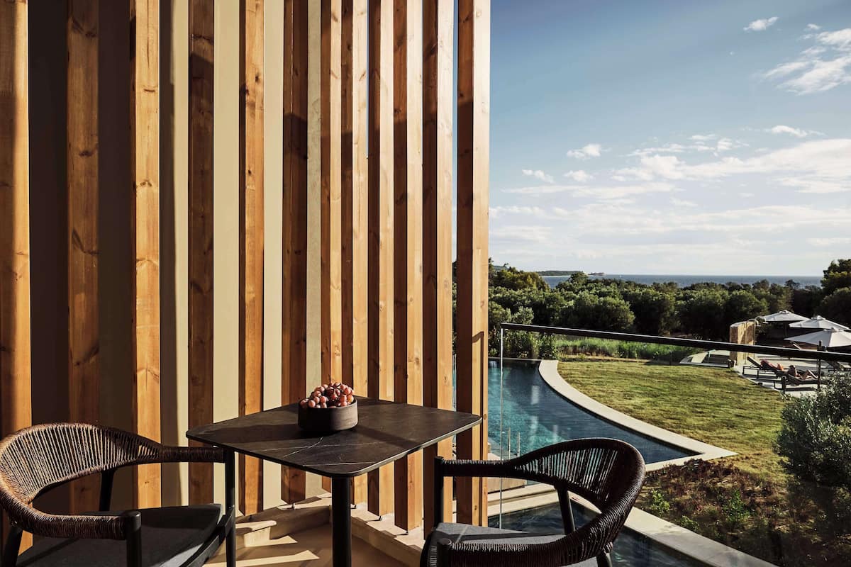 A wooden balcony with a table and chairs, overlooking a swimming pool and a landscaped garden.