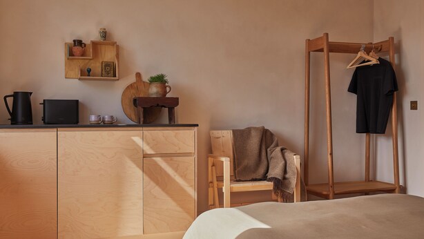 A minimalist bedroom with a wooden bed, a wooden dresser, a wooden chair, a wooden shelf with decorative items, and a wooden hanger rack with a black shirt. - Yarn - Elmo Luxury Double Suite (Ballycastle)