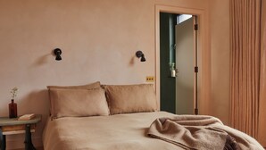 A bedroom with a bed, two wall-mounted light fixtures, a bedside table with a book and a vase, and a door leading to another room. - Yarn - Elmo Luxury Double Suite (Ballycastle)