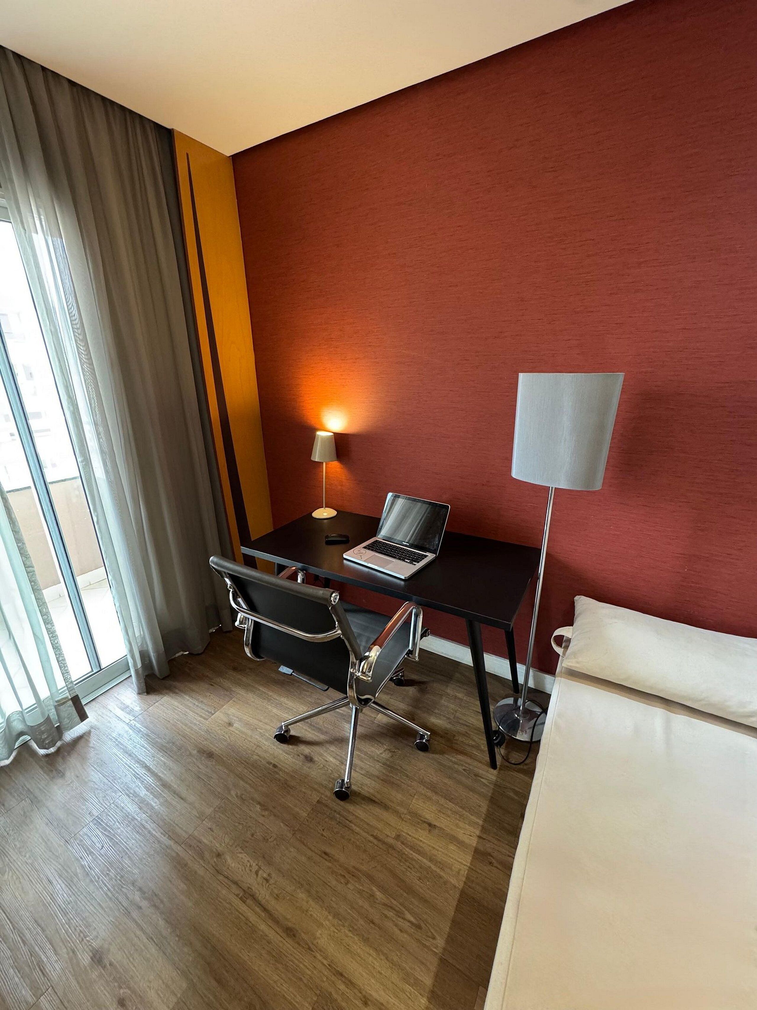 Une chambre d’hôtel avec un plancher en bois, un bureau avec un ordinateur portable, une chaise, une lampe sur pied et un lit.
