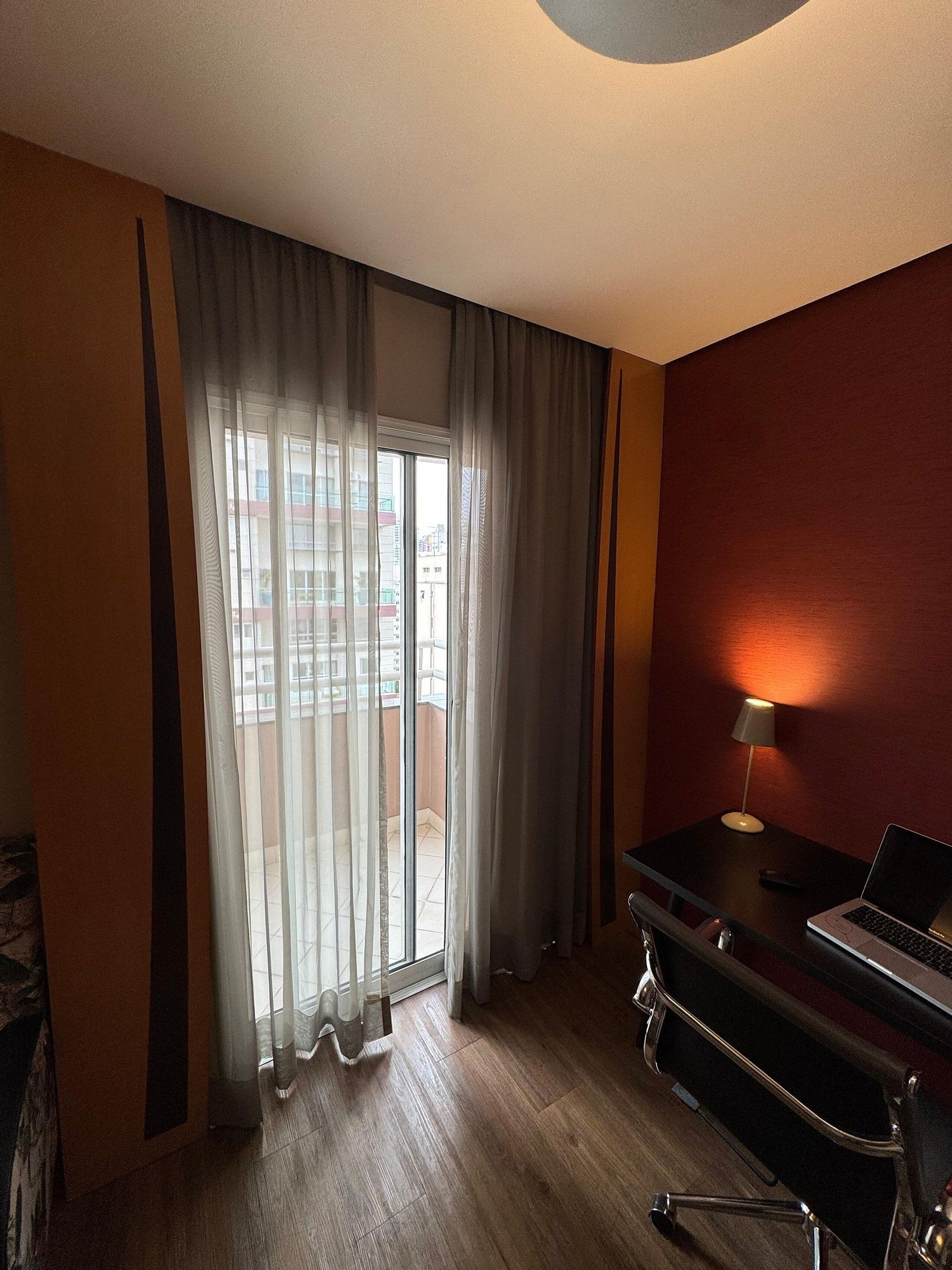 A hotel room with a large window, a desk with a laptop, and a standing lamp.