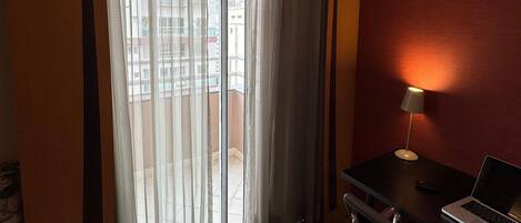 A hotel room with a large window, a desk with a laptop, and a standing lamp.