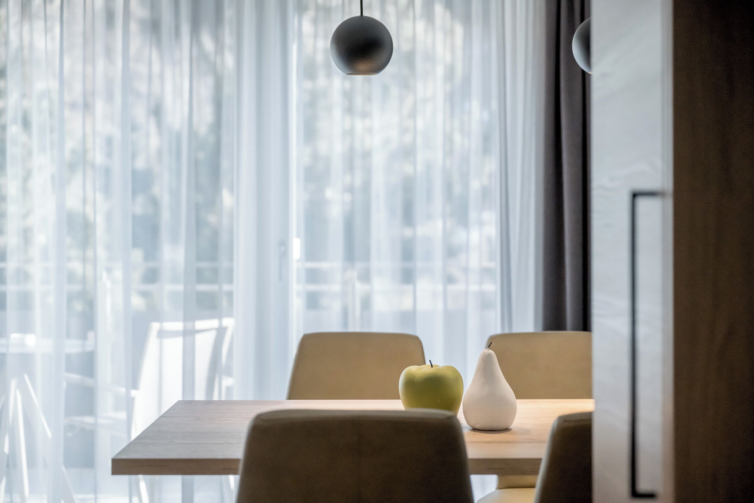 A dining area with a table, chairs, a hanging light fixture, and a view through sheer curtains.