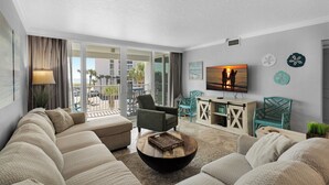 Una sala de estar moderna con un amplio sofá, una mesa de centro y un televisor. Tiene un balcón con vistas a palmeras y un edificio.