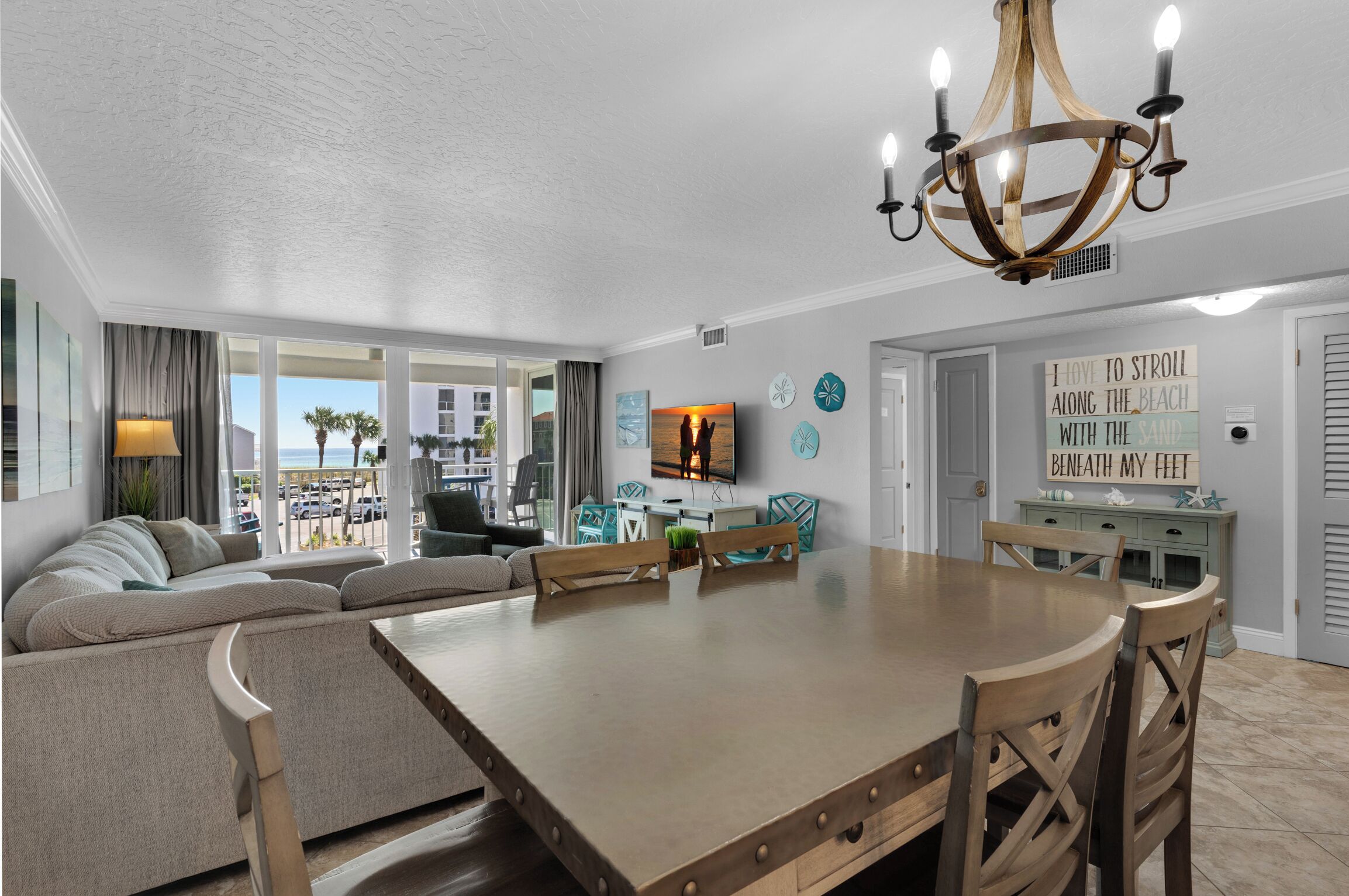 A dining area with a large wooden table and chairs, a chandelier, and a view of the beach through large windows.