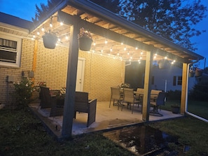 Une terrasse couverte, aménagée avec du mobilier d’extérieur, des guirlandes lumineuses et un mur en briques.