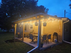 Een overdekt terras buiten, met een bakstenen muur, lichtjes en tuinmeubilair.