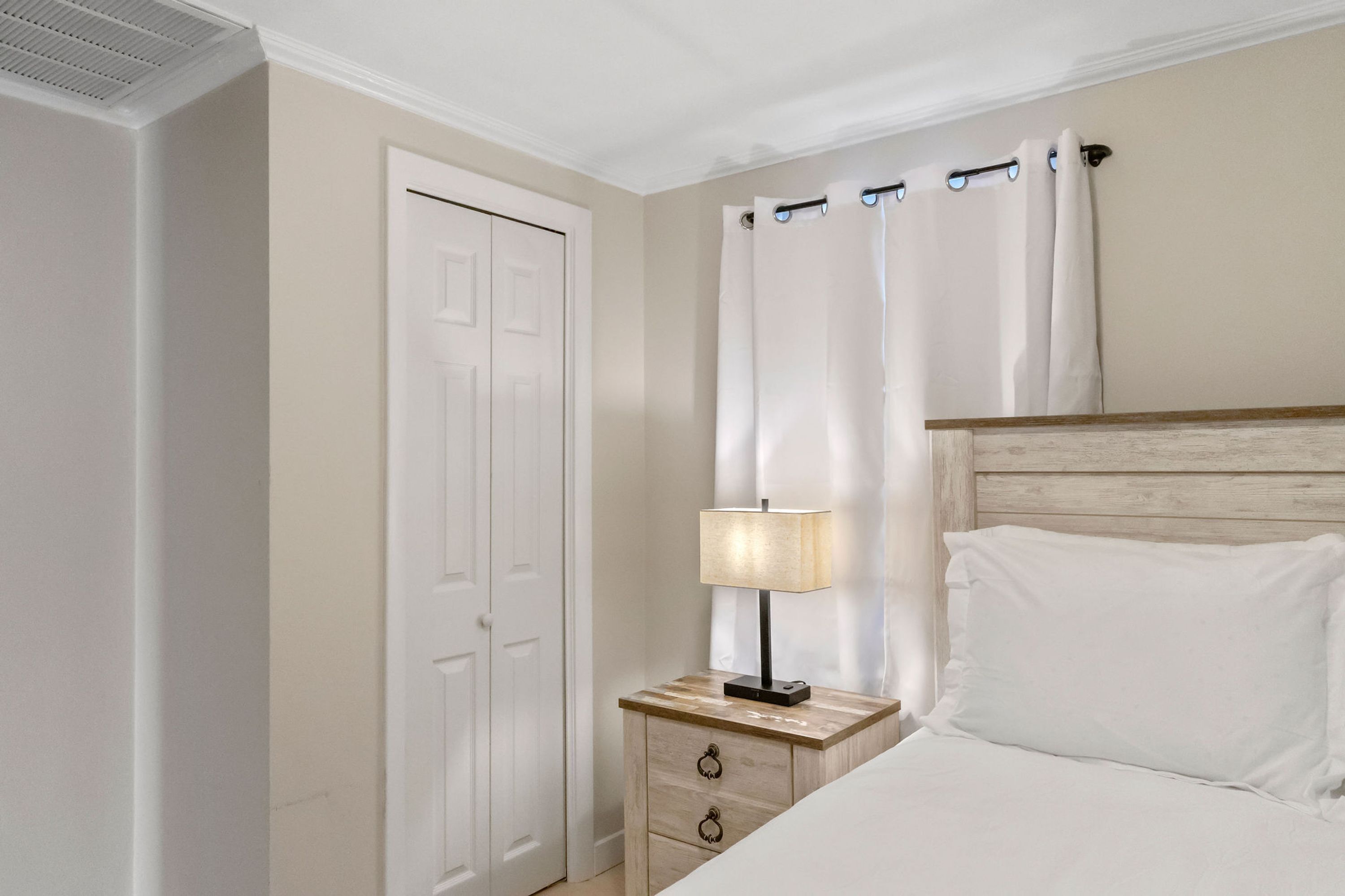 A bedroom with a bed, a nightstand with a lamp, a wooden headboard, a white door, and a window with curtains.