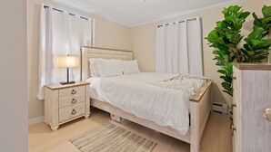 Un dormitorio con cama de madera, ropa de cama blanca, una mesita de noche con lámpara y una planta verde grande.