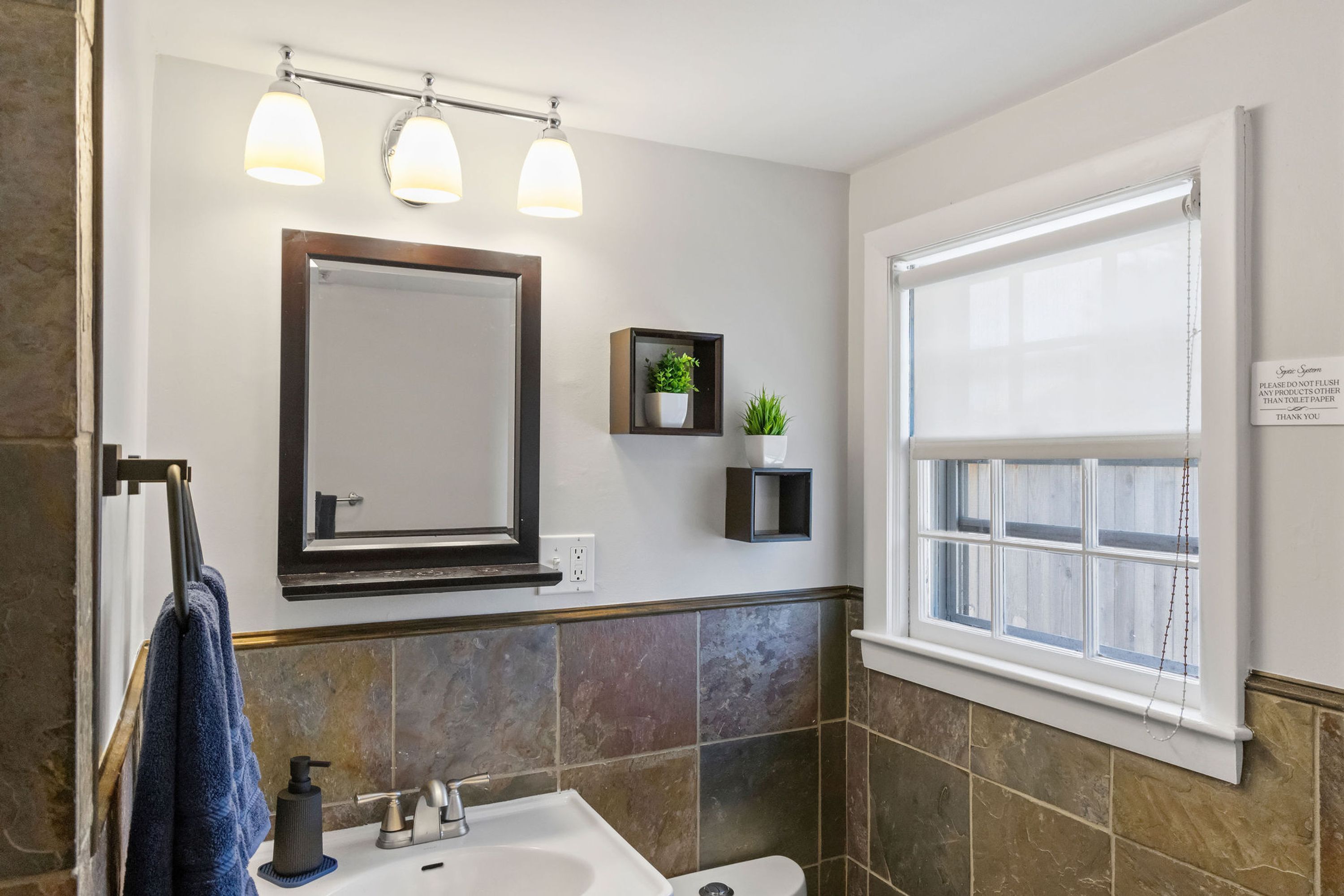 A bathroom with a large mirror, three wall-mounted lights, a sink, a toilet, and a window with a view.