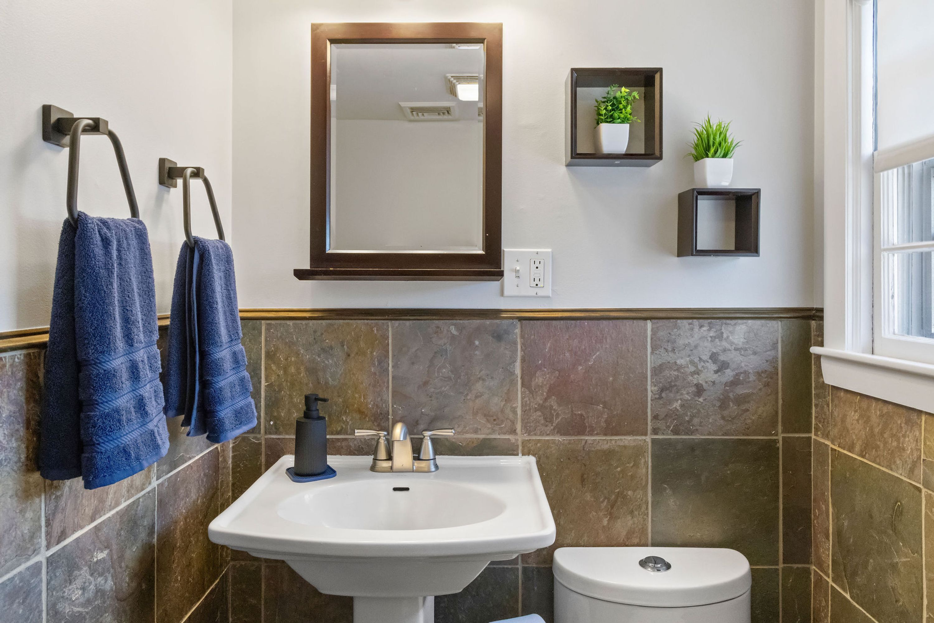 Une salle de bain avec un lavabo blanc, des toilettes et deux serviettes suspendues à un porte-serviettes. Il y a un miroir au-dessus du lavabo et une petite étagère avec une plante.