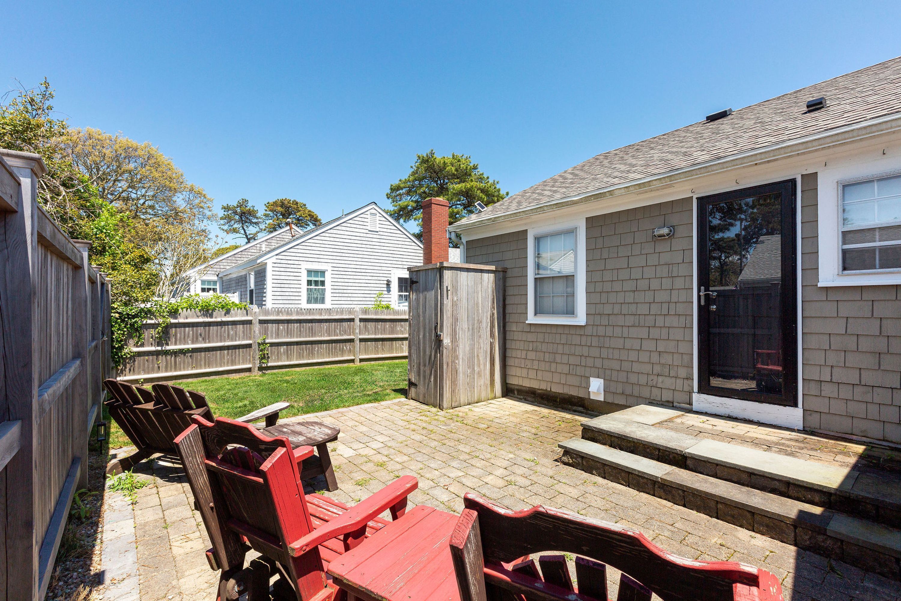 A backyard patio with a brick floor, wooden furniture, and a house in the background.
