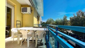 En balkon med et bord og stole, et blåt gelænder og udsigt til træer og parkerede biler.