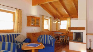 A cozy living room with a sofa, armchair, and dining table. A fireplace is visible in the corner, and wooden beams are on the ceiling. - Holiday House with Mountain View (Brixen im Thale)