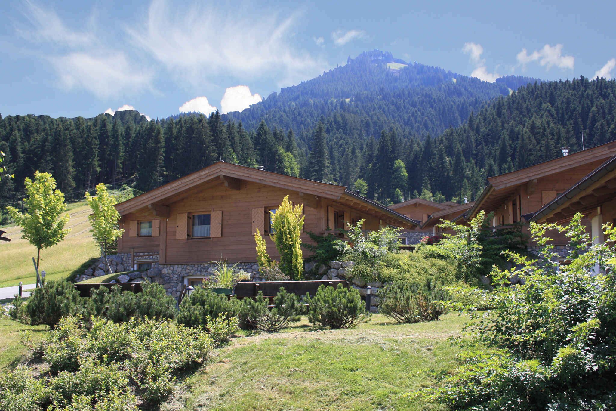 A wooden cabin with a stone foundation, surrounded by lush greenery and a mountainous backdrop.