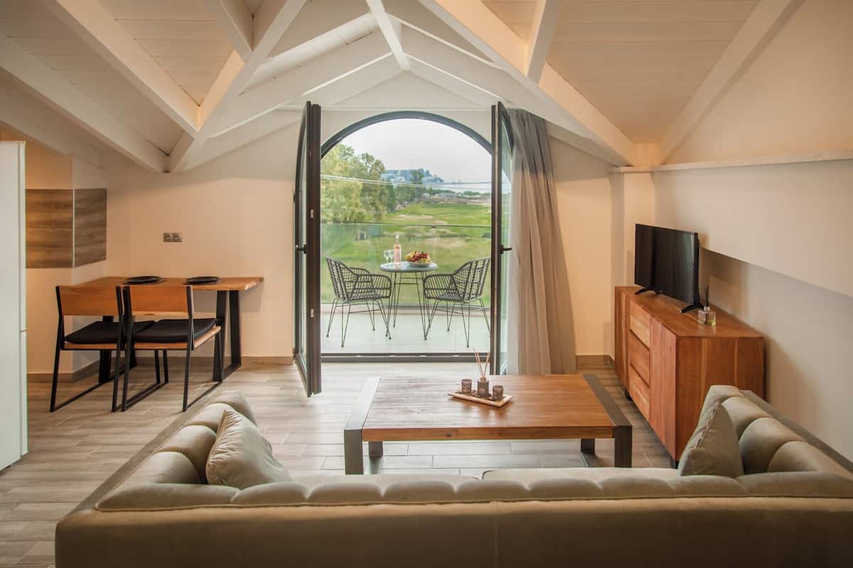 Un salon moderne avec un grand canapé, une table basse en bois, un coin repas avec des chaises, un téléviseur posé sur un meuble en bois et une vue sur un terrain de golf, visible à travers une porte vitrée.