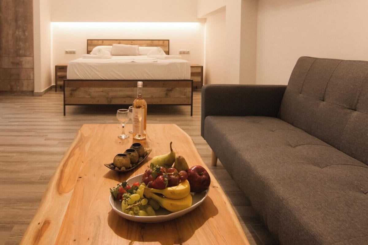 Une chambre d’hôtel moderne avec une table à manger en bois, agrémentée d’un plateau de fruits et d’une bouteille, un canapé gris et un lit recouvert de draps blancs.