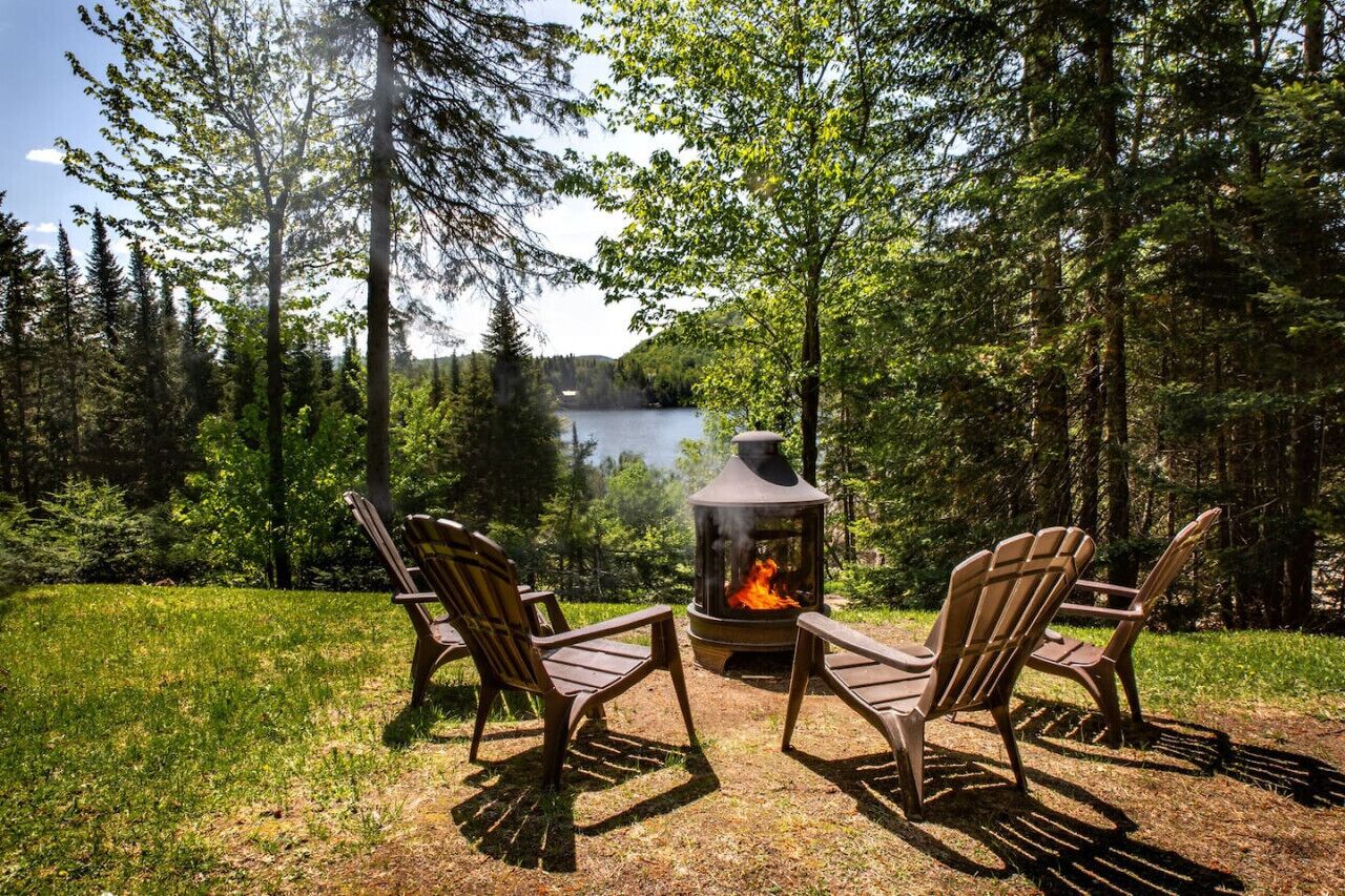 Zona boscosa con un hogar para fogatas y dos sillas Adirondack mirando un lago.