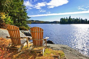 Kaksi Adirondack-tyylistä puutarhatuolia järven rannalla, jonka reunalla on metsä.