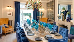 Une salle à manger élégante, avec une longue table dressée pour un repas, des chaises à revêtement bleu, un lustre et une cheminée.