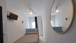 A narrow hotel room with a single bed, a ceiling fan, a mounted TV, and a large mirror on the wall.