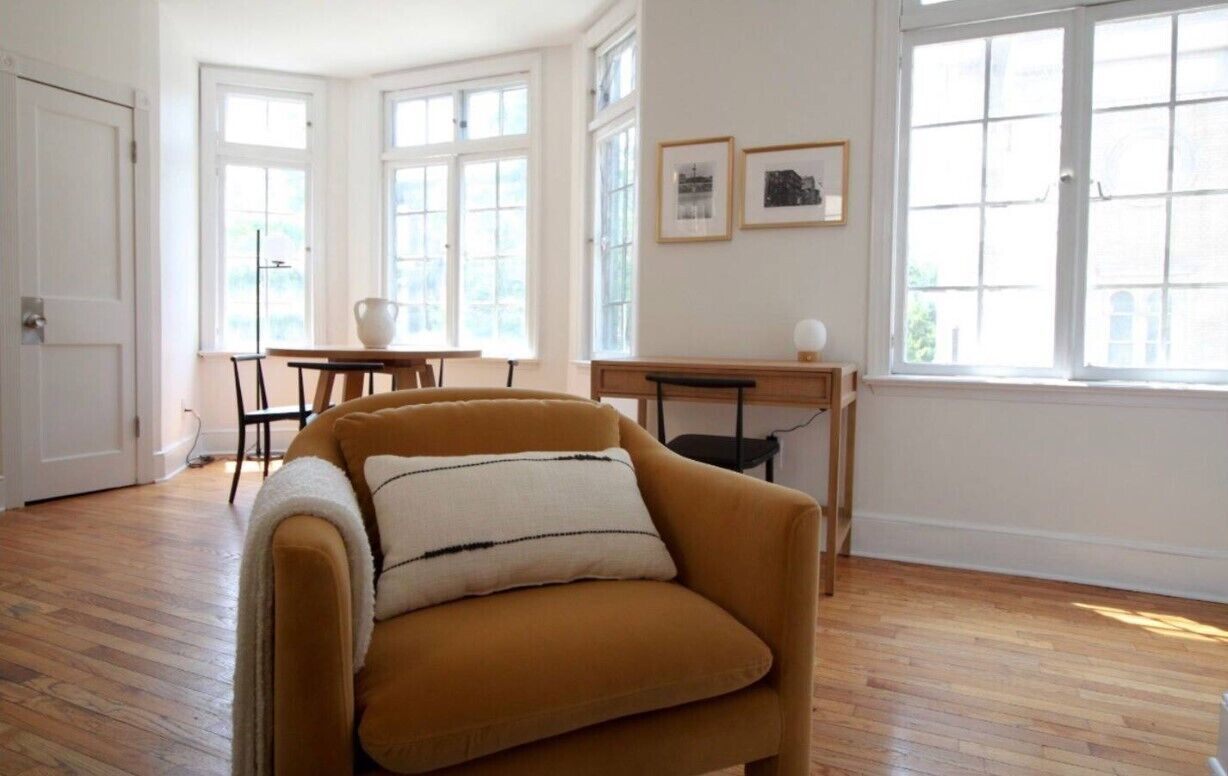 A room with a wooden floor, a sofa with a white cushion, a table, chairs, and large windows.