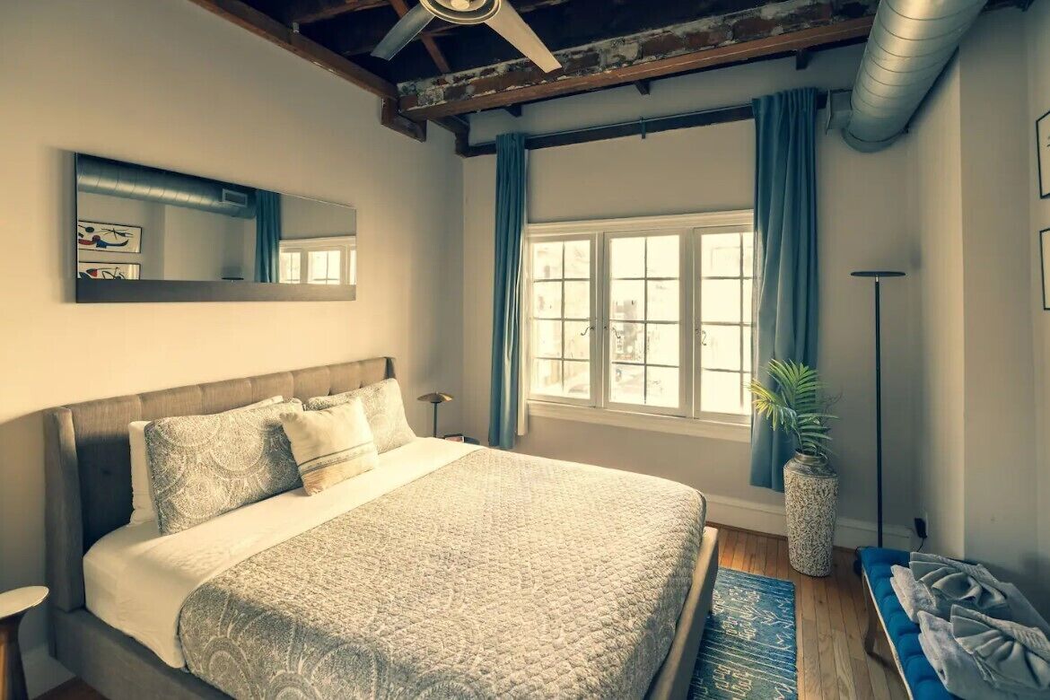 A bedroom with a large bed, a ceiling fan, a window with curtains, a potted plant, and a blue bench.