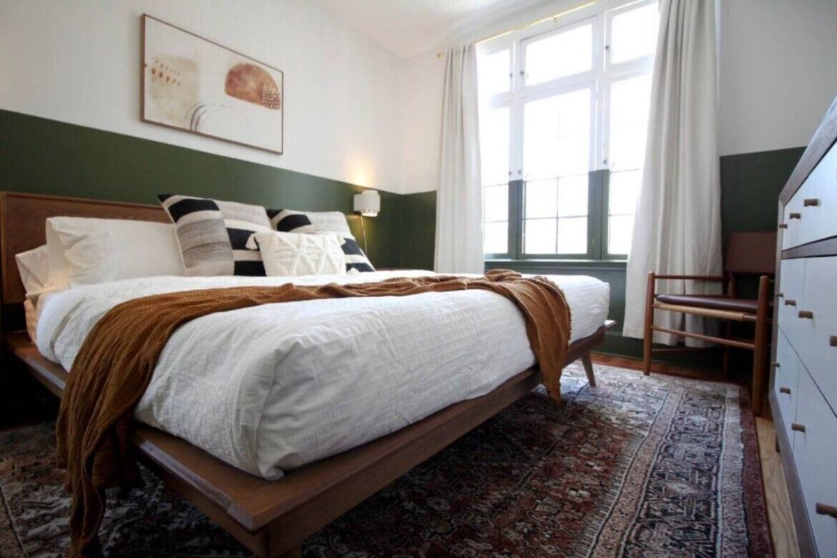 Une chambre à coucher comprenant un lit, une table de chevet, une commode, une fenêtre avec des rideaux et une lampe fixée au mur.