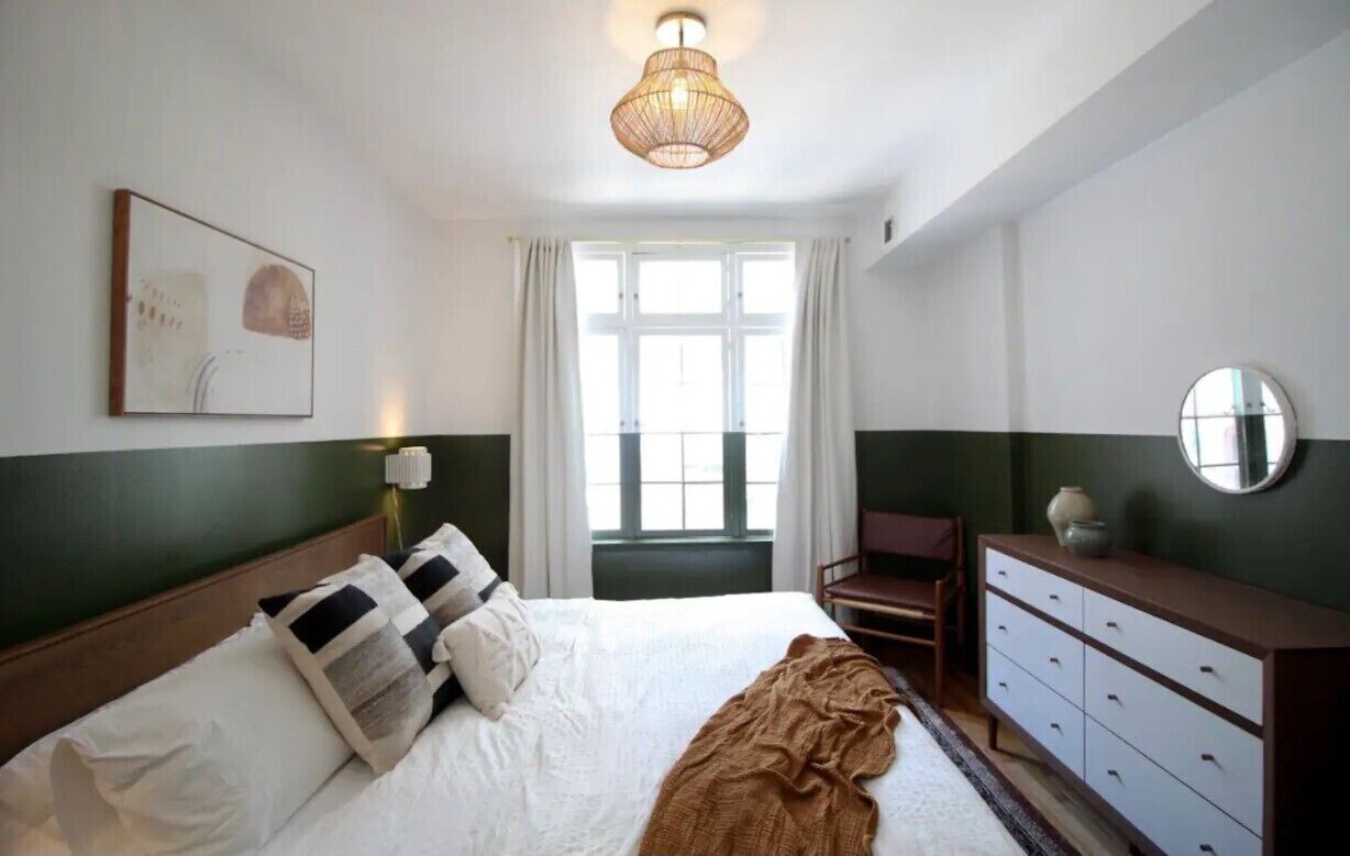 A bedroom with a bed, a dresser, a chair, a window with curtains, and a hanging light fixture.