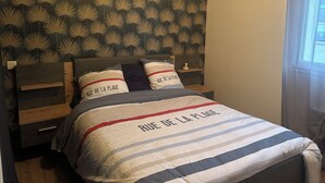 A bed with a striped bedspread and pillows with text, a headboard with a leaf pattern, and a nightstand with a small vase. - Au délices d'Anne Laure !   (Pleumeur-Gautier)