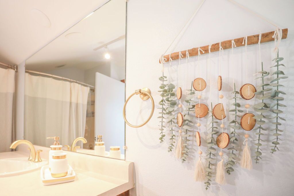 A bathroom with a large mirror, gold faucets, and a wall-mounted wooden decoration with hanging elements.
