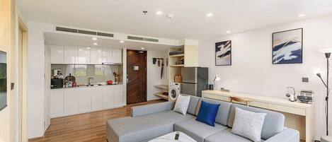 A modern living room with a grey sofa, a white coffee table, and a kitchen area with white cabinets and a wooden door.