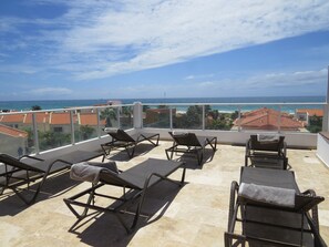 Une terrasse sur le toit, dotée de quatre chaises longues, d’une balustrade en verre et offrant une vue sur la mer et les bâtiments.