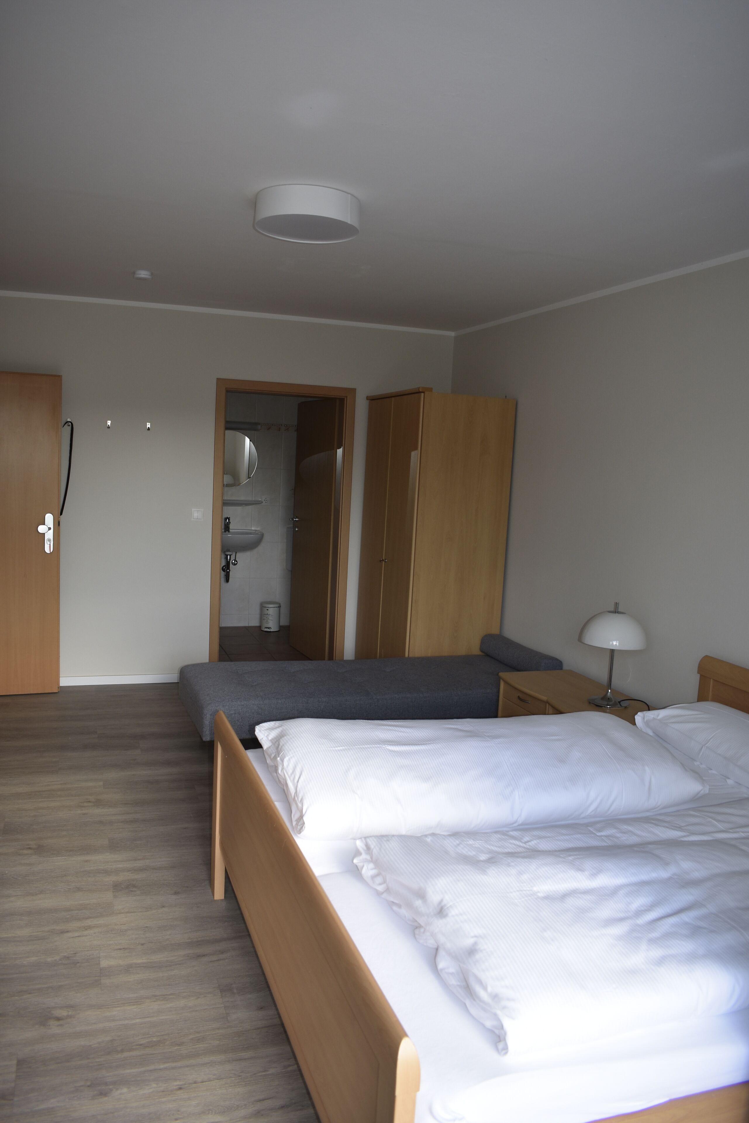 Un dormitorio con dos camas, un armario de madera, una mesita de noche con lámpara y un baño visible a través de una puerta abierta.