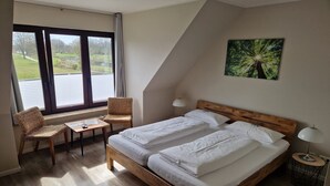 A bedroom with a wooden bed, two bedside tables, a chair, a small table, and a window with blinds. - Pension zur Schleuse (Witzeeze)