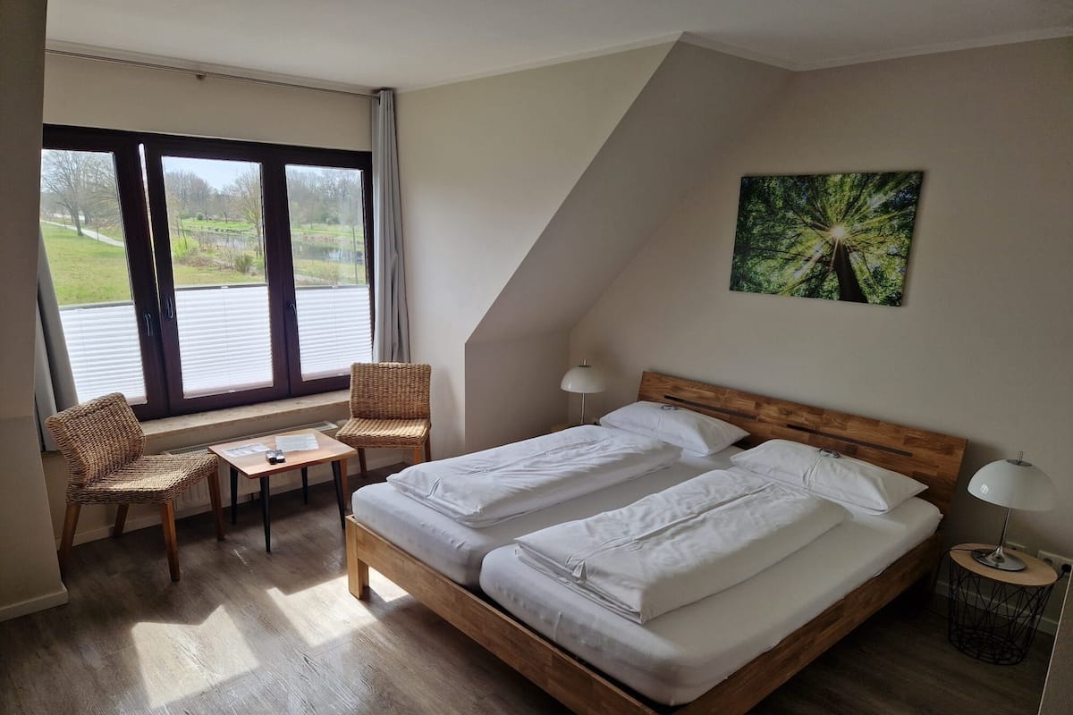 A bedroom with a wooden bed, two bedside tables, a chair, a small table, and a window with blinds.