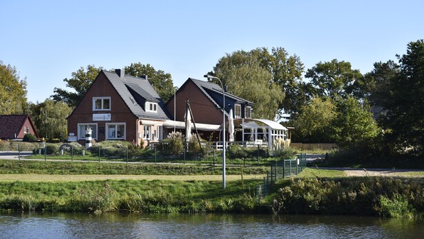 A riverside building with a brick facade, a covered outdoor area, and a grassy bank. - Pension zur Schleuse (Witzeeze)