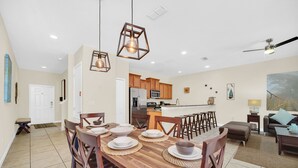 A dining area with a wooden table set for four, a kitchen in the background, and a ceiling fan.