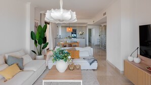 Amplia sala de estar con un sofá blanco, una mesa de centro y una planta grande. Se ve una cocina al fondo con una mesa de comedor y sillas.