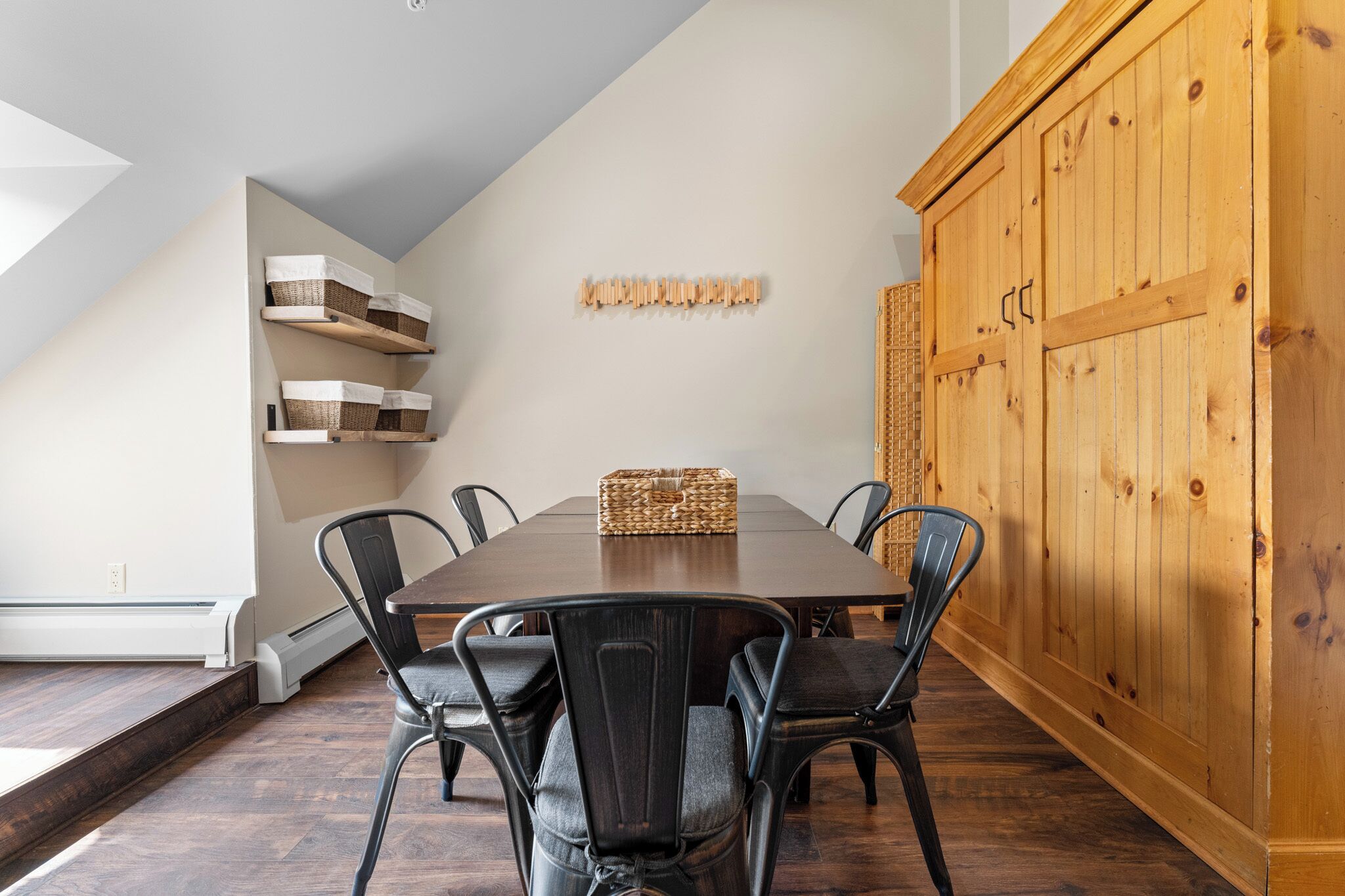 A dining area with a wooden table, six chairs, a cabinet, and shelves with baskets.