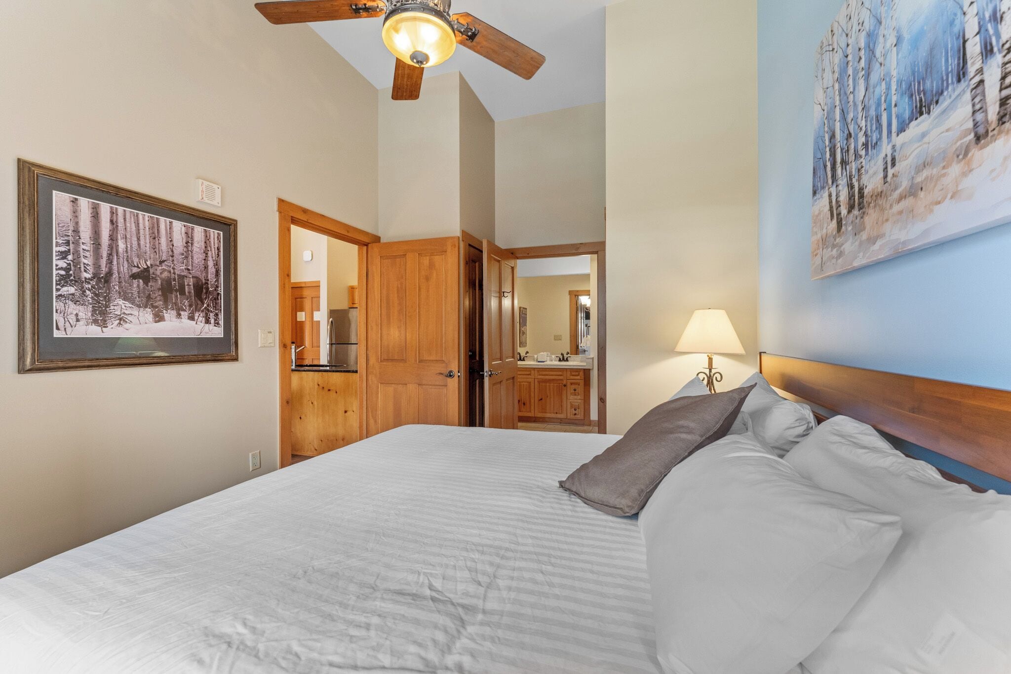 A bedroom with a bed, a ceiling fan, a painting of a forest, and a bathroom visible through an open door.