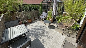 Een terras met een zithoek, inclusief een tafel en stoelen, omringd door plantenbakken en een glazen overkapping.