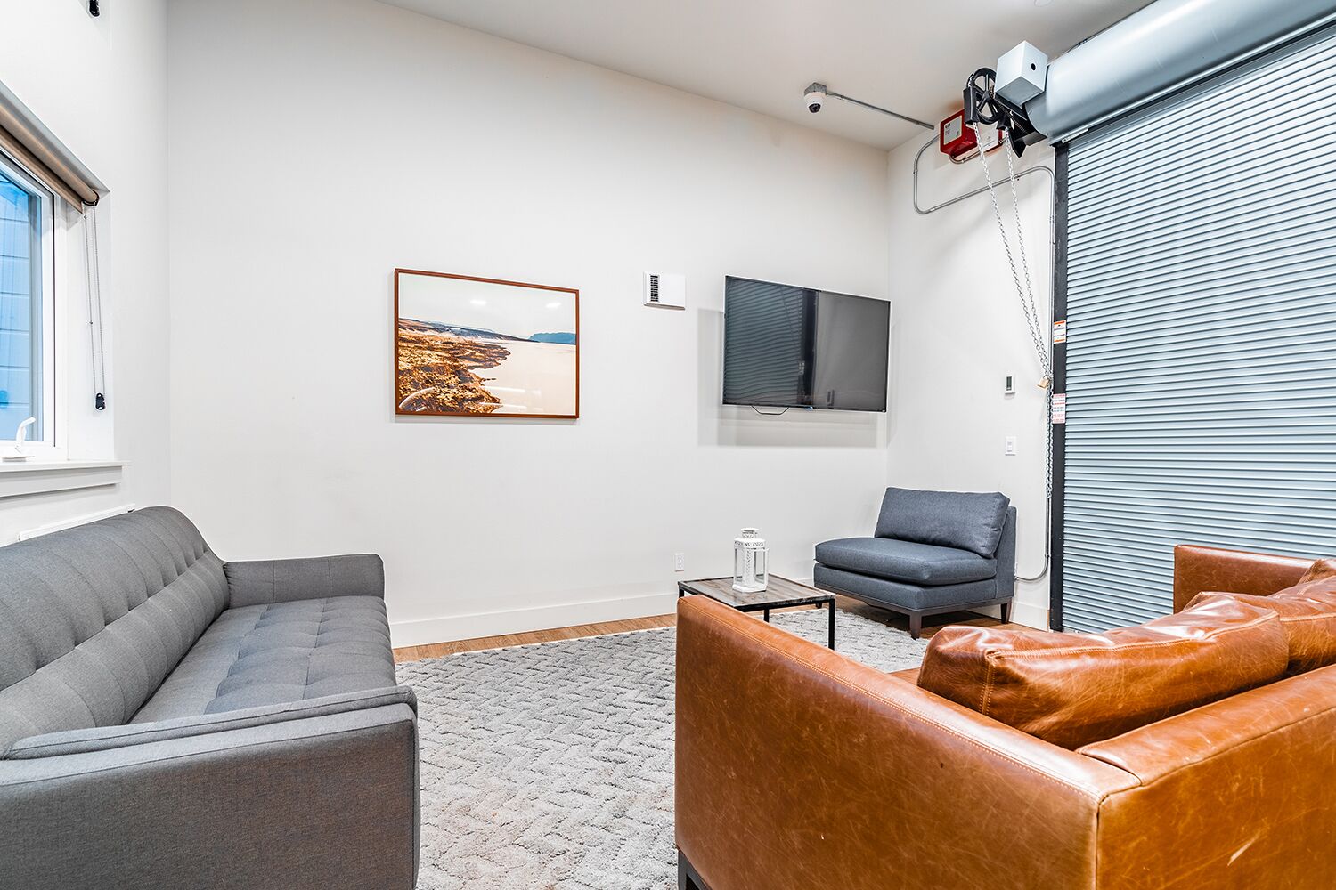 A modern living room with a grey sofa, a brown leather couch, a grey armchair, a flat-screen TV, and a framed landscape picture on the wall.