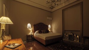 Une chambre d’hôtel avec un grand lit, deux lampes de chevet, une table en bois avec un livre, un lustre et un tapis décoratif.