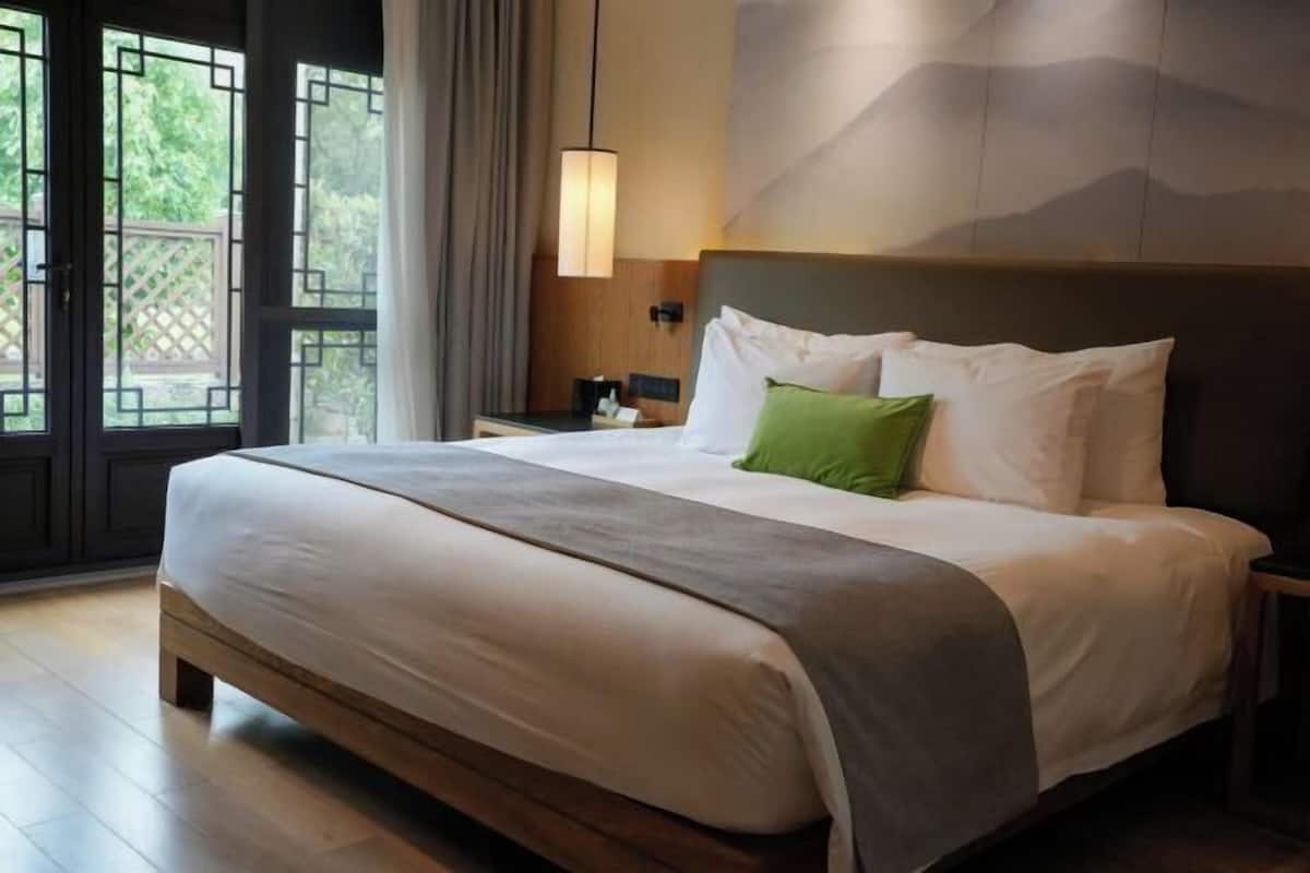 A hotel room with a large bed, a nightstand, a lamp, and a view of greenery through the window.