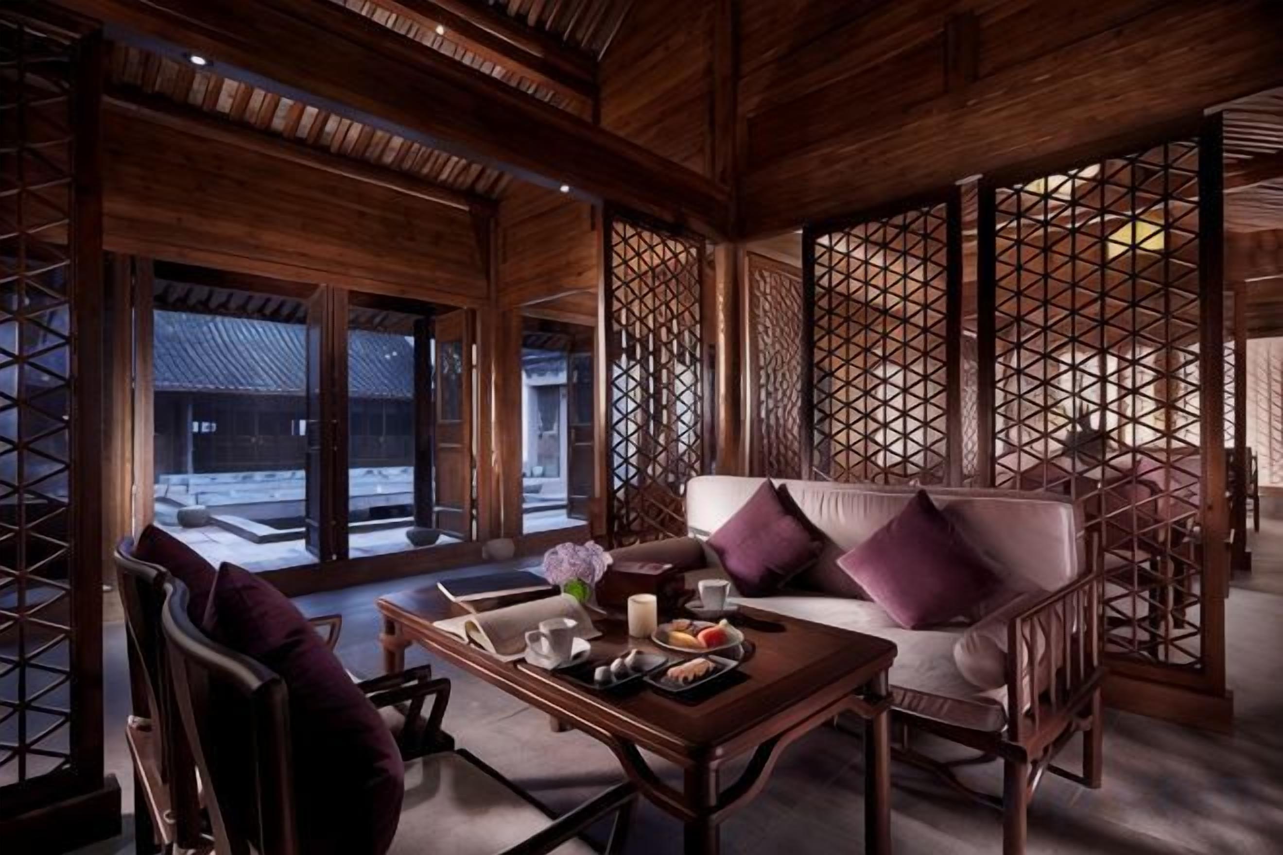 A traditional wooden interior with a sofa and a table set with food and drinks, large windows, and a view of the outside.