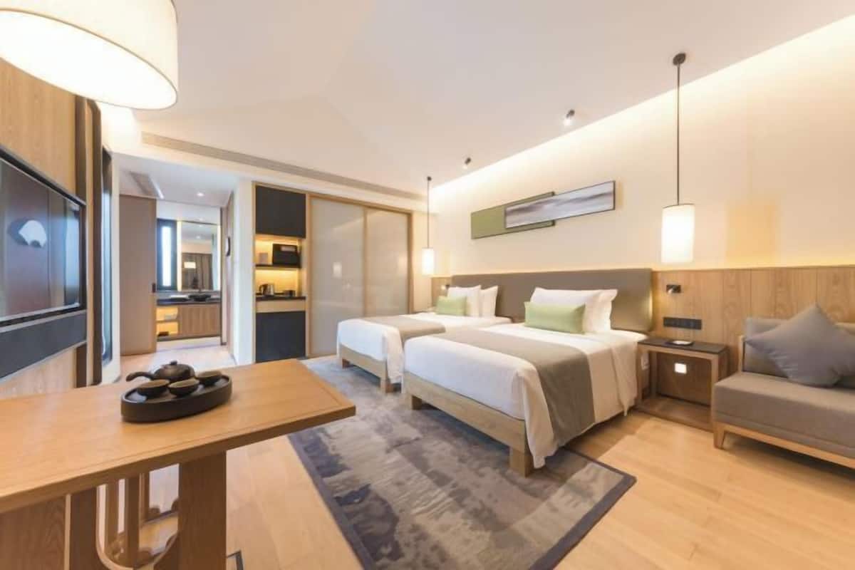 A hotel room with two beds, a desk, a sofa, a dining table, and a kitchen area visible through an open door.
