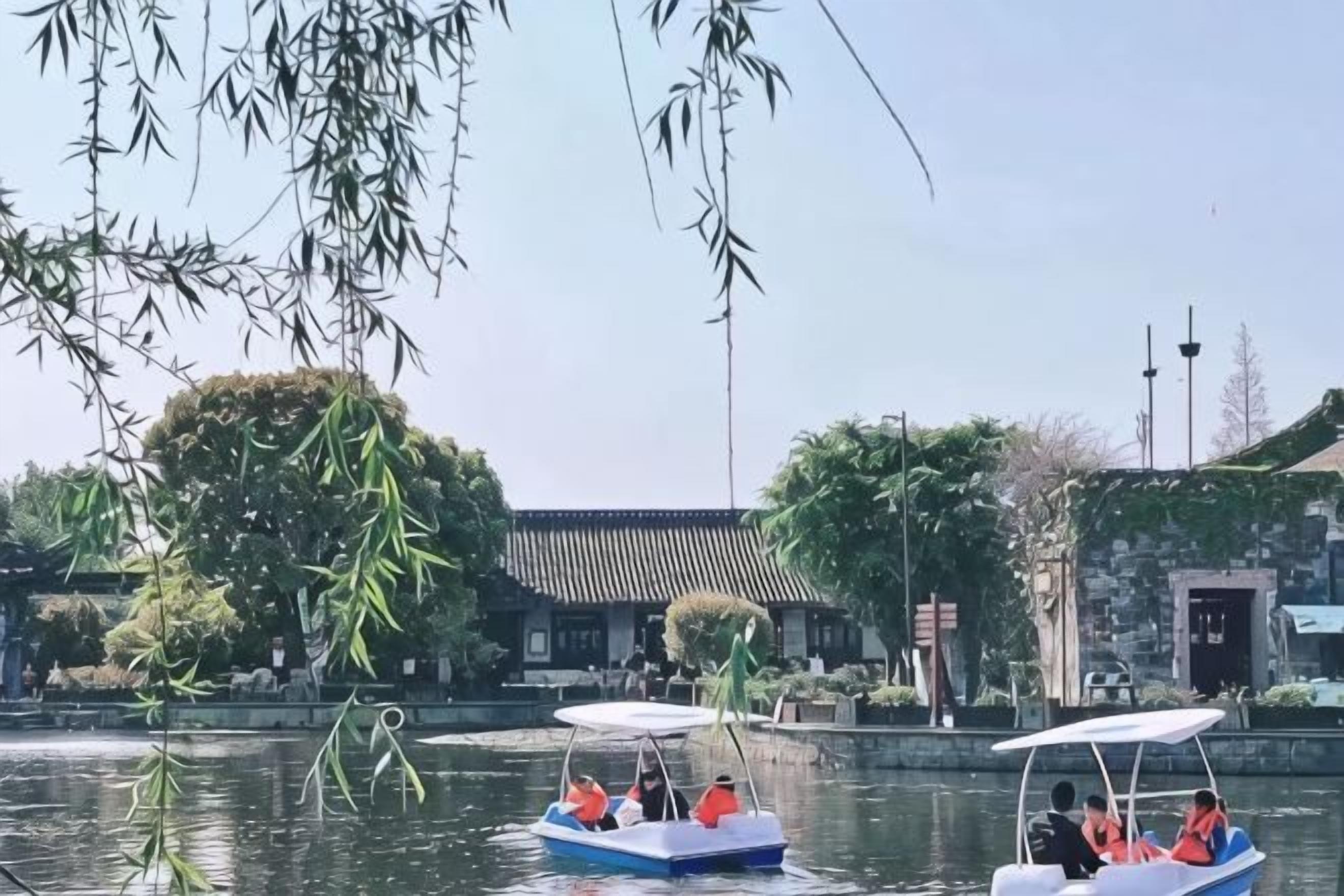 Un bâtiment traditionnel avec un toit en tuiles, un plan d’eau où flottent des bateaux et un arbre aux branches pendantes.